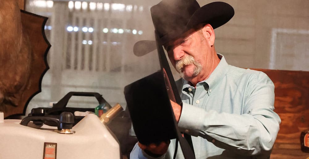 Un vaquero con bigote gris sosteniendo un sombrero negro sin forma y colocándolo en el vapor.